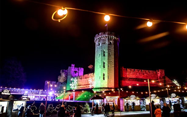 Warwick Castle illuminated at night during the Light Trail event, with visitors exploring festive stalls.