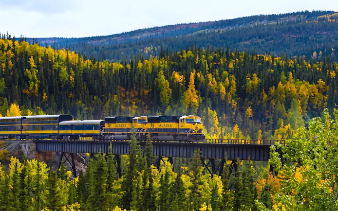 Alaska Railroad train crossing a bridge through autumn forest scenery.