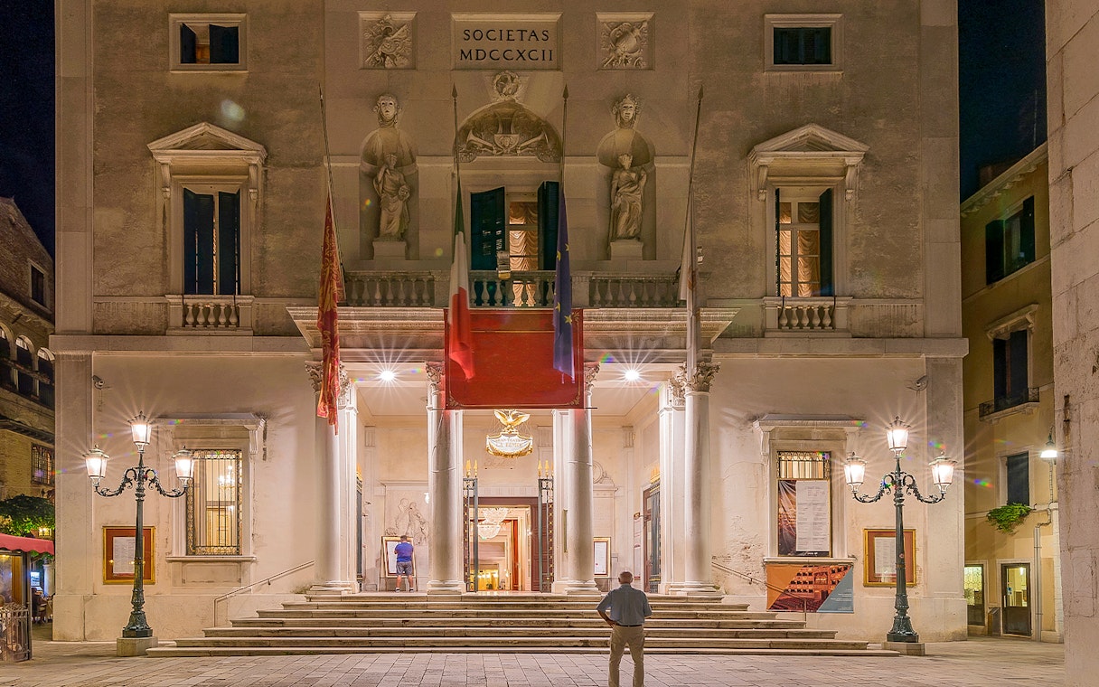 Facade of Fenice Theater in Venice at night, featuring illuminated entrance and architectural details.