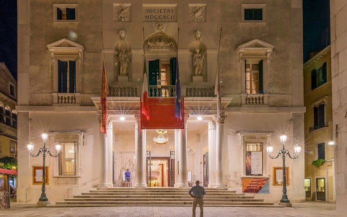 Facade of Fenice Theater in Venice at night, featuring illuminated entrance and architectural details.