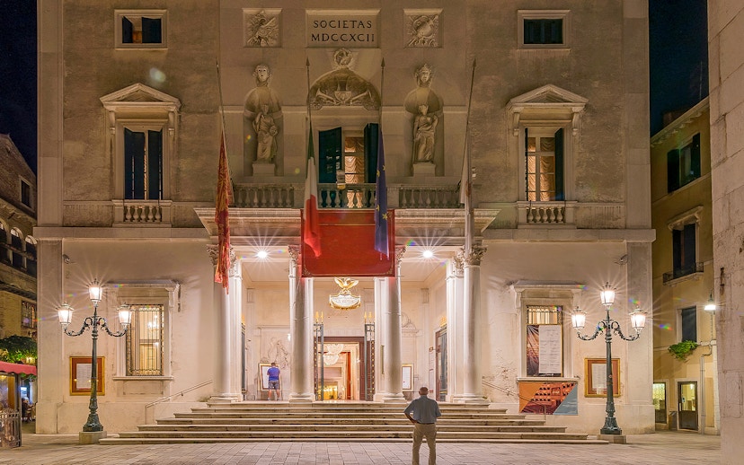 Facade of Fenice Theater in Venice at night, featuring illuminated entrance and architectural details.