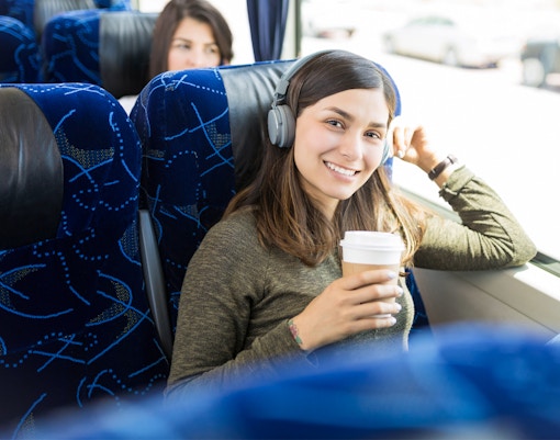 Woman sitting on a blue chair ina shuttle bus, listening to music and posing for the camera while holding a cup of coffee in one hand.