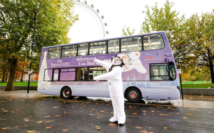 Snowman character in front of The Snowman™ Afternoon Tea Bus in London.