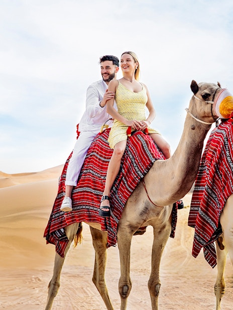 Couple riding camels in desert, part of Hot Air Balloon Experience with Vintage Land Rover Drive.