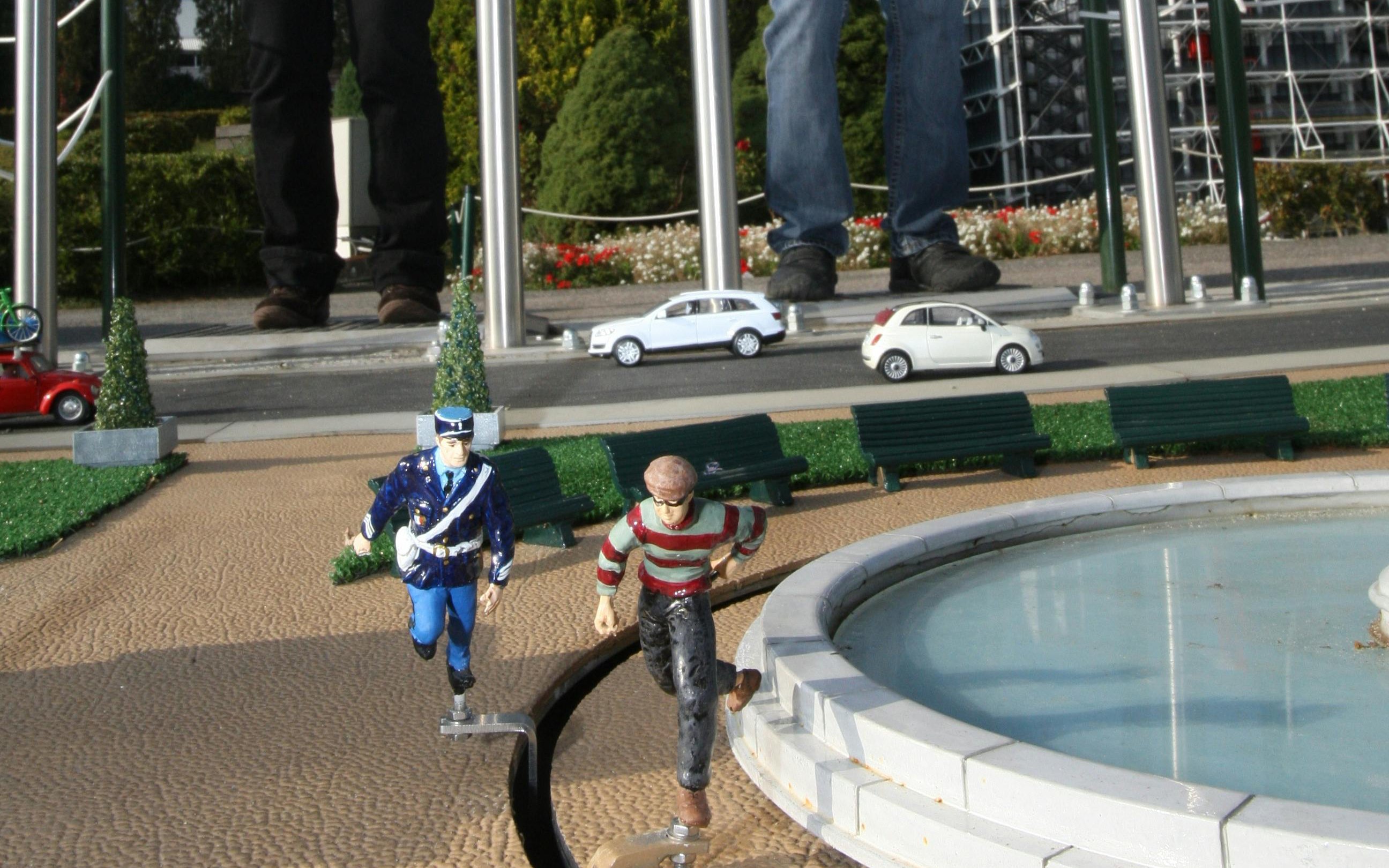 Miniature figures in a chase scene at Mini-Europe, Brussels, with tiny cars and benches nearby.