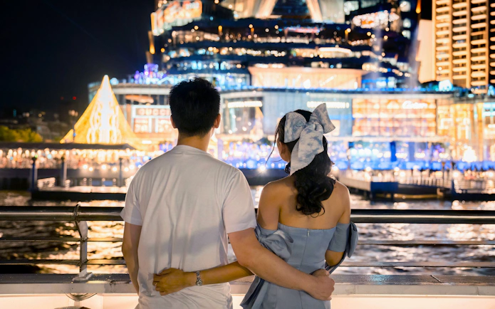 Young couple embracing on cruise ship deck, viewing light show at department store, White Orchid River Cruise.