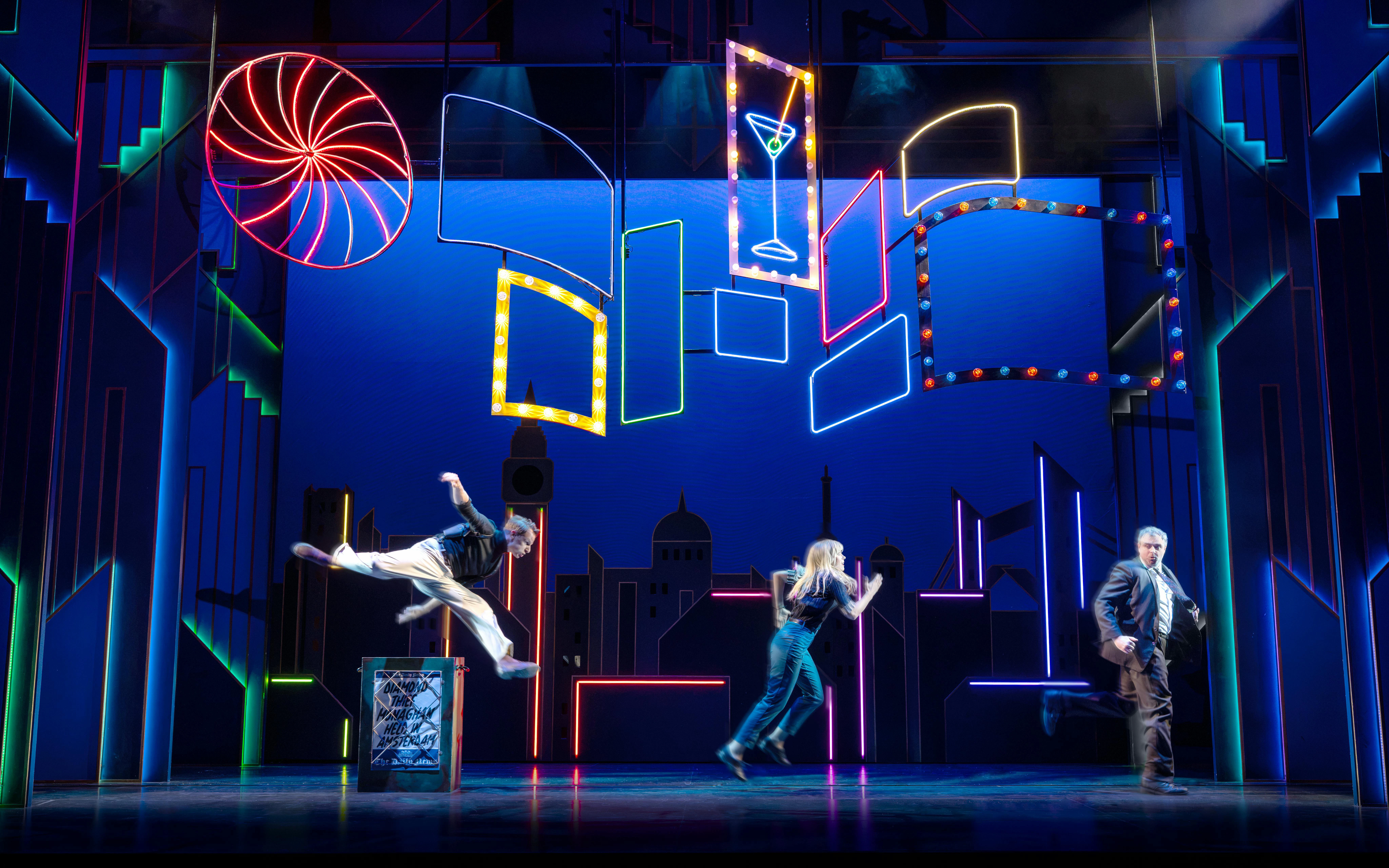 Actors performing a scene from "The Comedy About Spies" with neon cityscape backdrop.