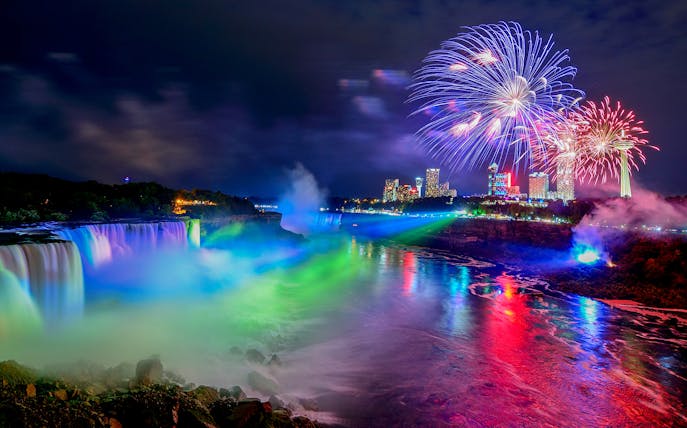 Niagara Falls illuminated at night with fireworks and city skyline in the background.