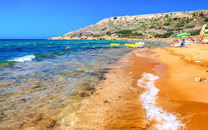 Golden sand and clear waters at Ramla Beach, Gozo, with lush green hills in the background.