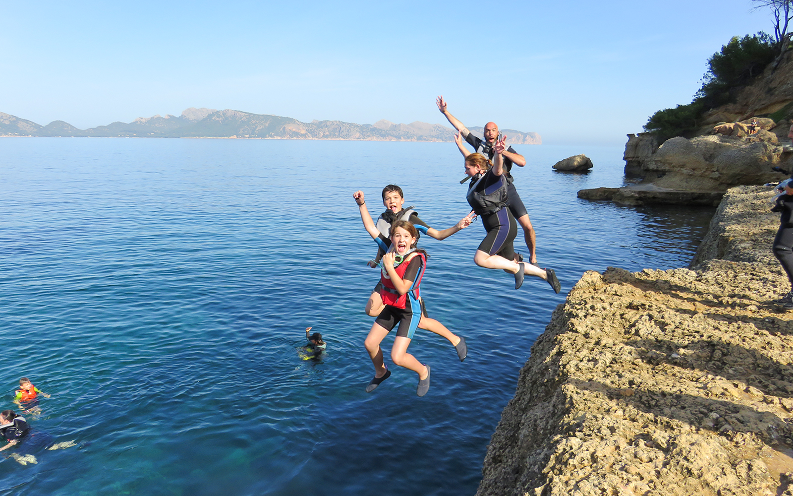 Group cliff jumping into the sea in Mallorca, with snorkelers and kayakers nearby.