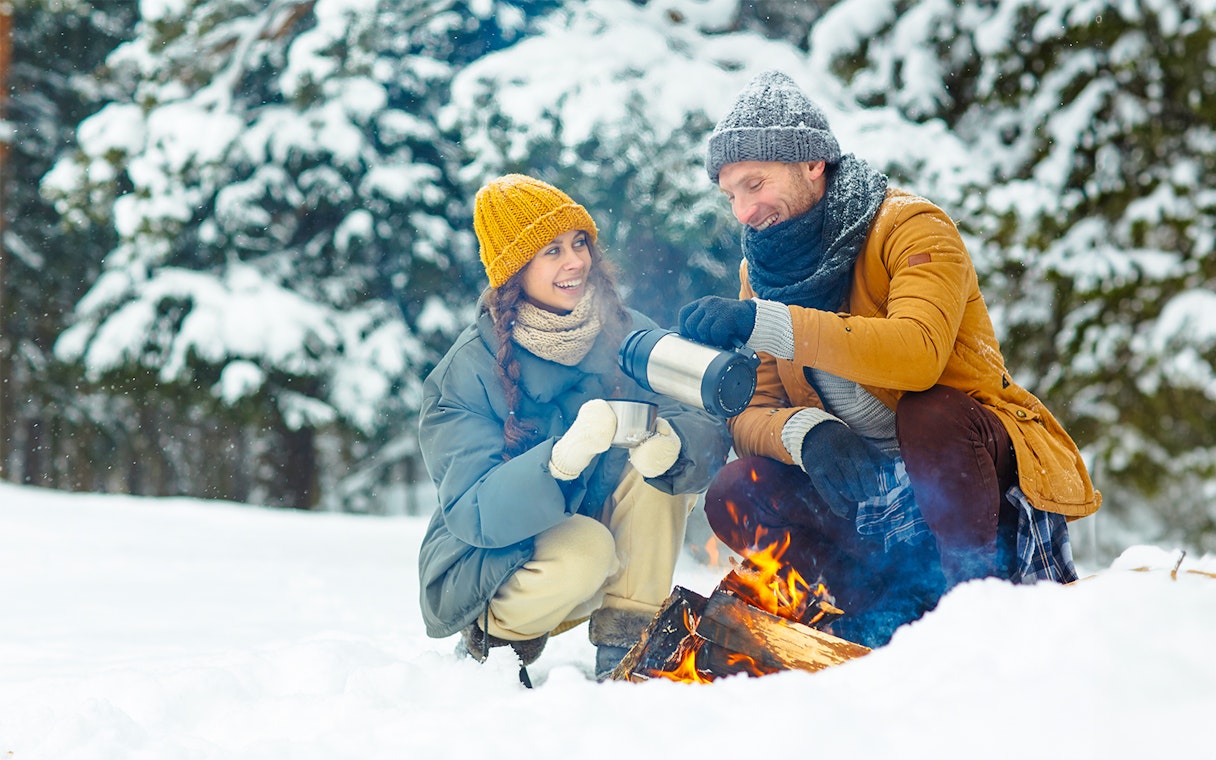 Couple enjoying hot drinks by a campfire on a snowy Northern Lights tour.