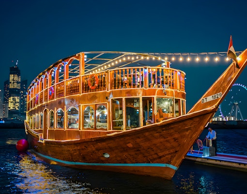 Illuminated dhow boat on Dubai Marina with city skyline and Ferris wheel in the background.
