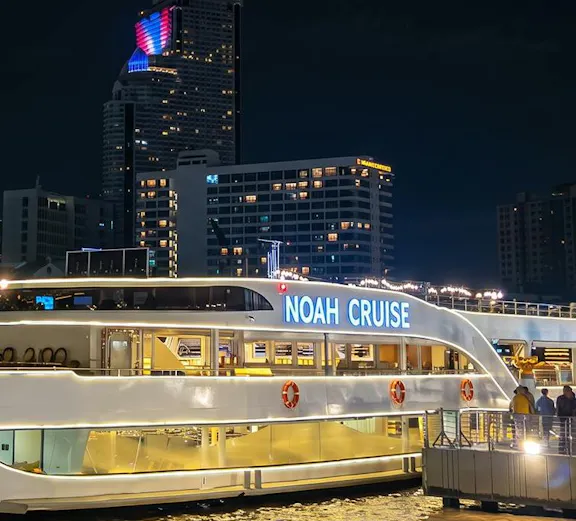 Noah Cruise ship docked at night in Bangkok with city skyline in the background.