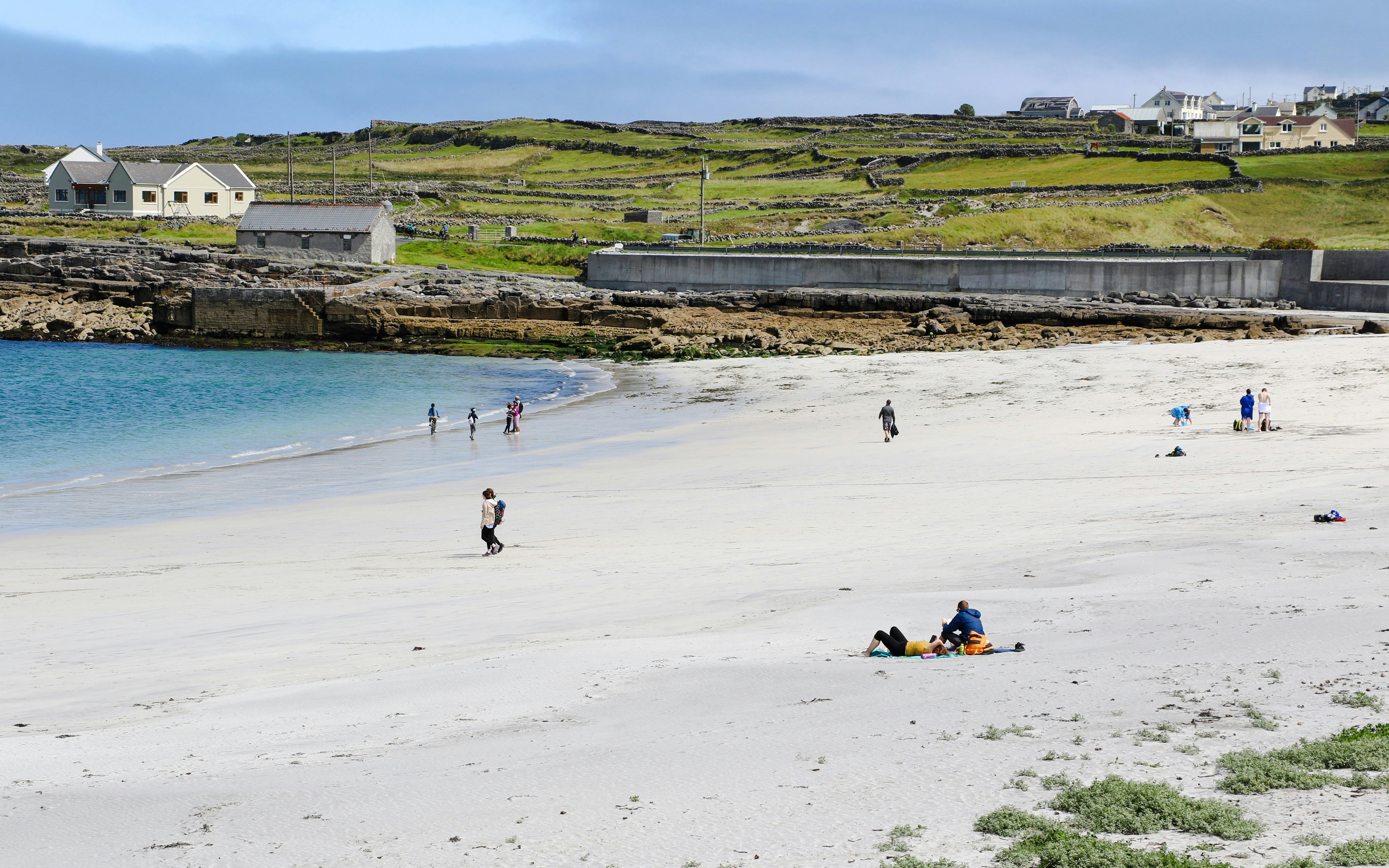 People walking and relaxing on Kilmurvey Beach, Aran Islands, Ireland, with stone houses in the background.