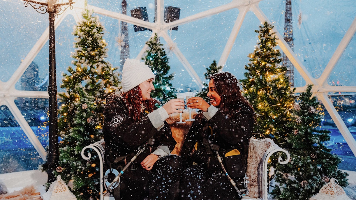 Guests toasting inside the Up at the O2 Snow Globe Experience with festive trees.