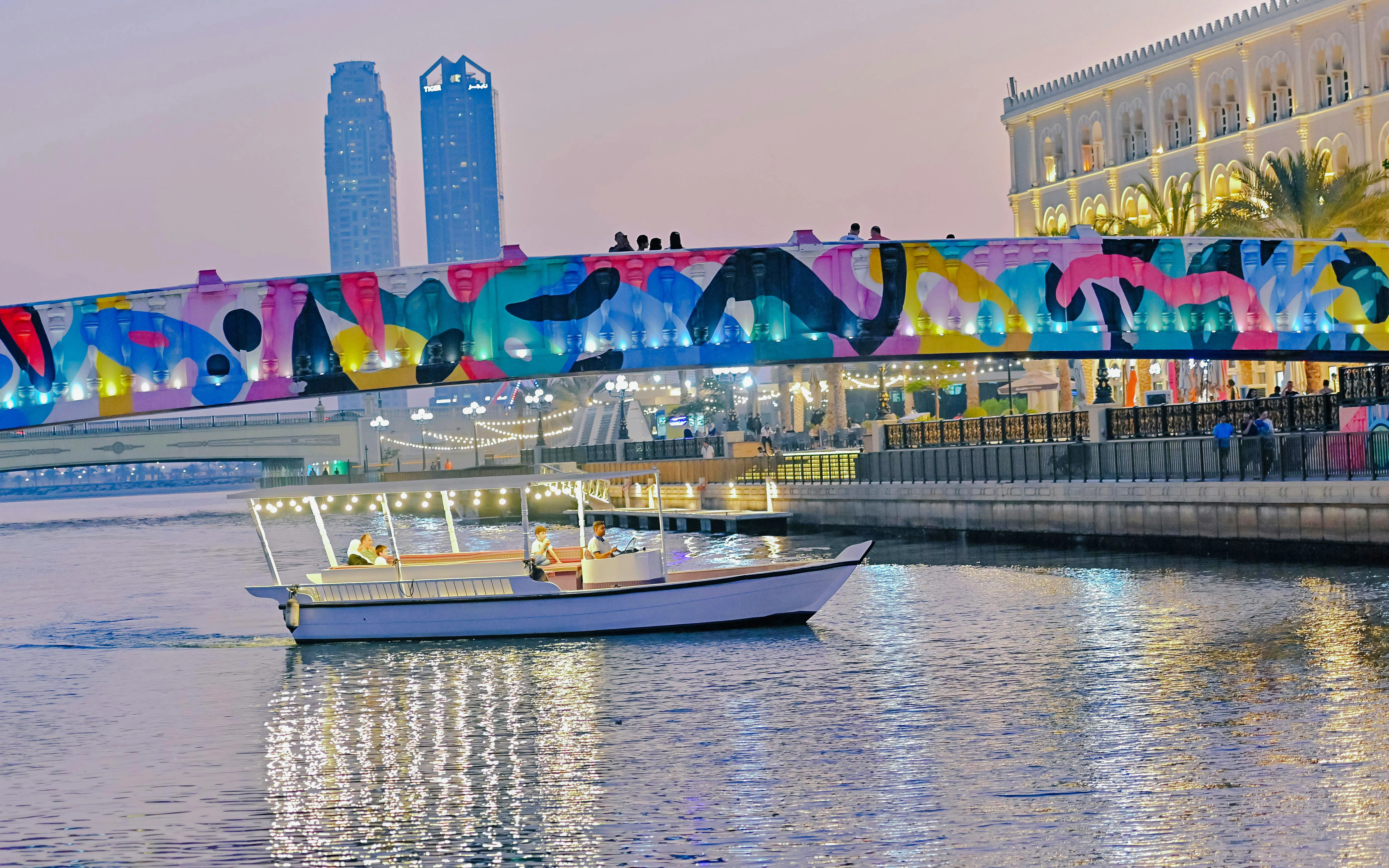 Boat cruising under colorful bridge in Sharjah during evening tour.