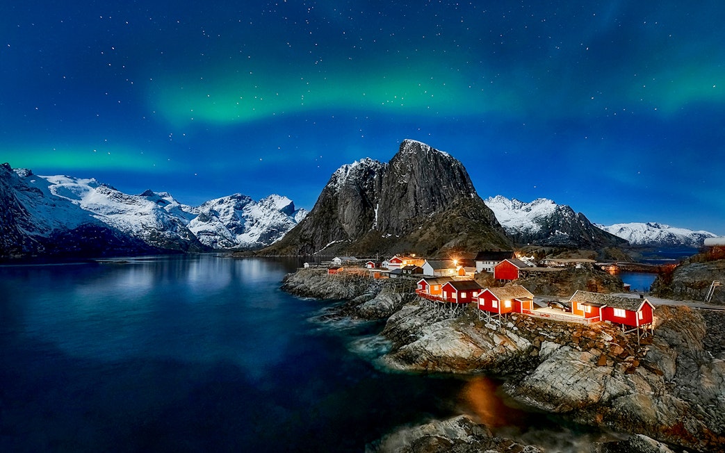 Northern Lights over snowy mountains and red cabins in Lofoten, Norway.