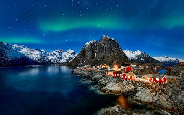 Northern Lights over snowy mountains and red cabins in Lofoten, Norway.