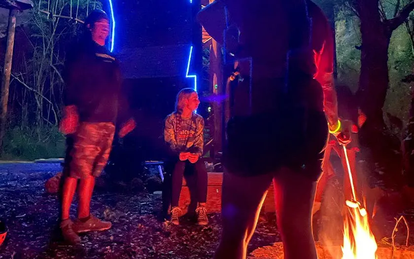 Campfire gathering during night zipline adventure with people roasting marshmallows.