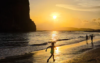Child running on Railay Beach at sunset, Krabi, with silhouettes of people and cliffs.