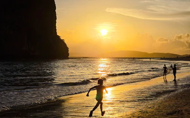 Child running on Railay Beach at sunset, Krabi, with silhouettes of people and cliffs.