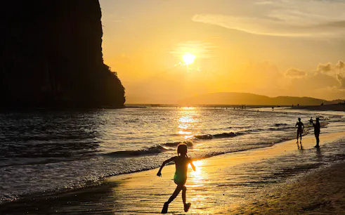 Child running on Railay Beach at sunset, Krabi, with silhouettes of people and cliffs.