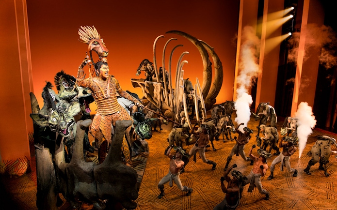 Stage performance of The Lion King with actors in elaborate costumes and set design.