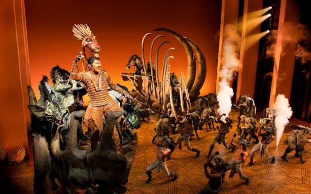 Stage performance of The Lion King with actors in elaborate costumes and set design.