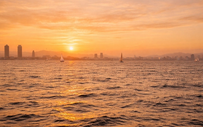 Yachts sailing at sunset with Barcelona skyline in the background.