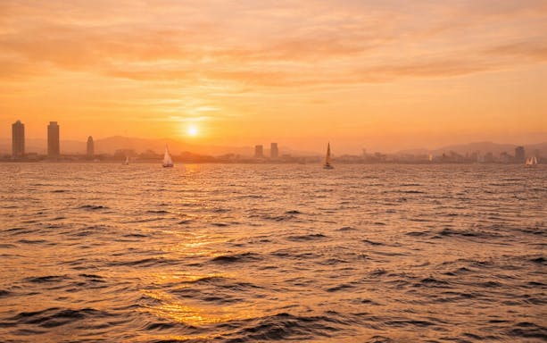 Yachts sailing at sunset with Barcelona skyline in the background.