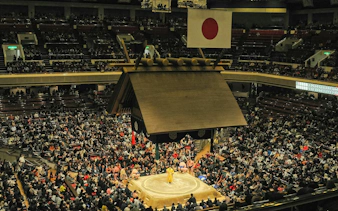Grand Sumo Tournament in Tokyo with a crowded stadium and sumo wrestlers in the ring.