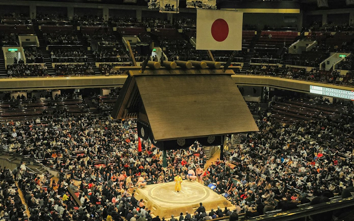 Grand Sumo Tournament in Tokyo with a crowded stadium and sumo wrestlers in the ring.