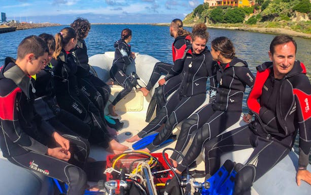 Group preparing for scuba diving baptism in Bosa Sea, wearing wetsuits on a boat.