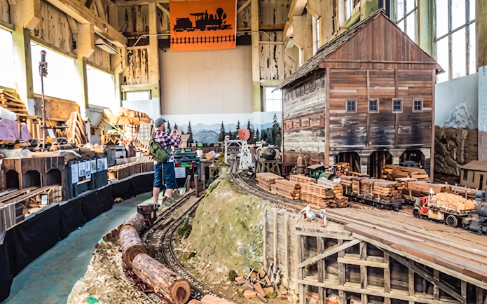 Visitor exploring model train exhibit with detailed railroads and wooden structures.