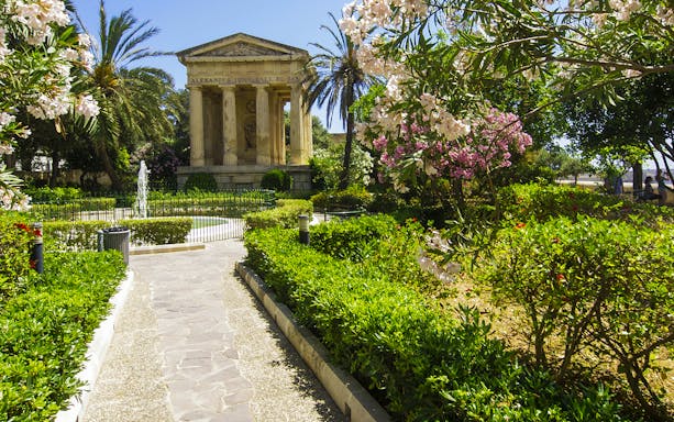 Pathway leading to Lower Barrakka Gardens monument in Valletta, surrounded by lush greenery.
