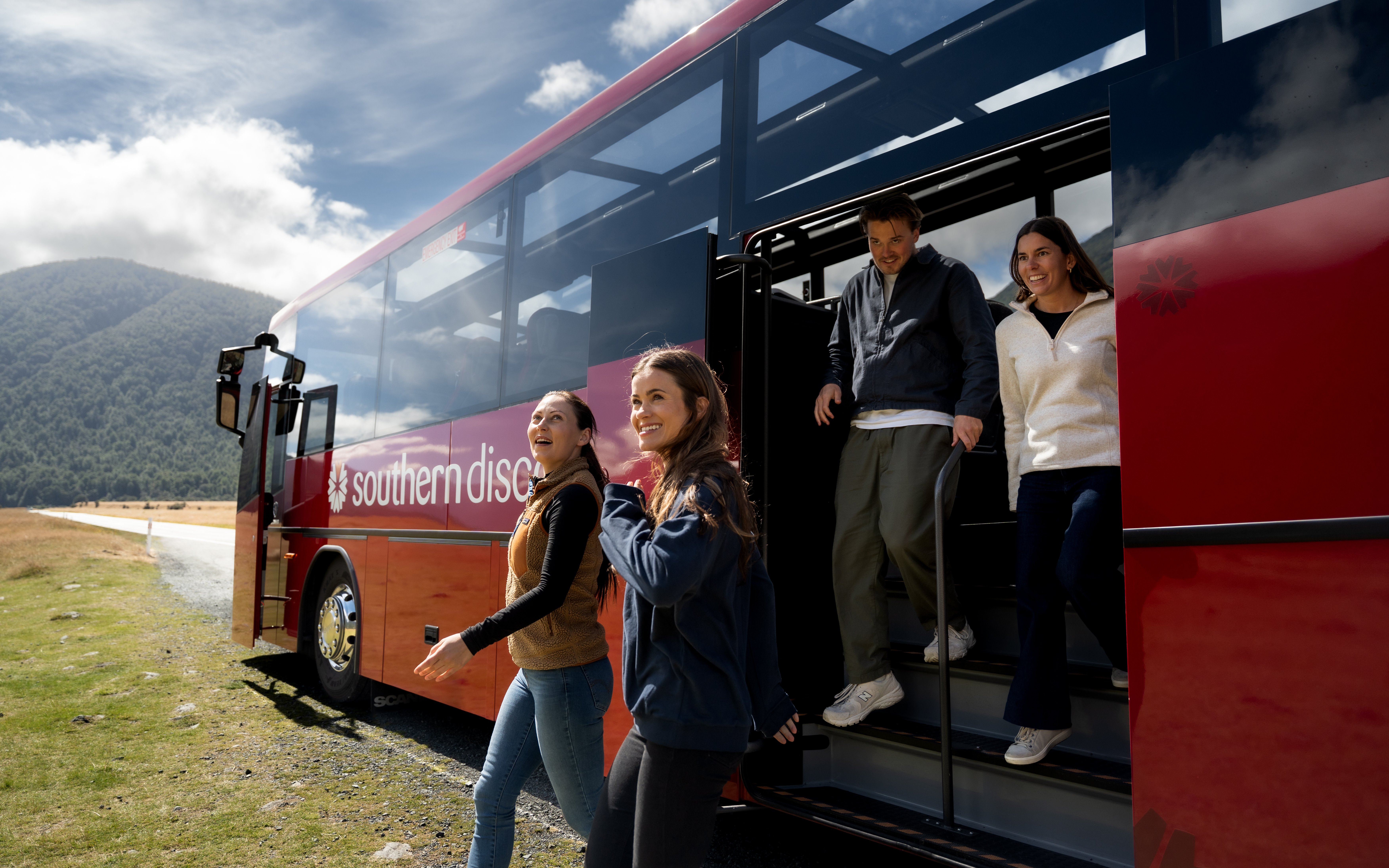 Tourists exiting a coach at Milford Sound with scenic mountain backdrop.