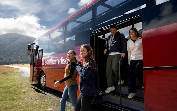 Tourists exiting a coach at Milford Sound with scenic mountain backdrop.