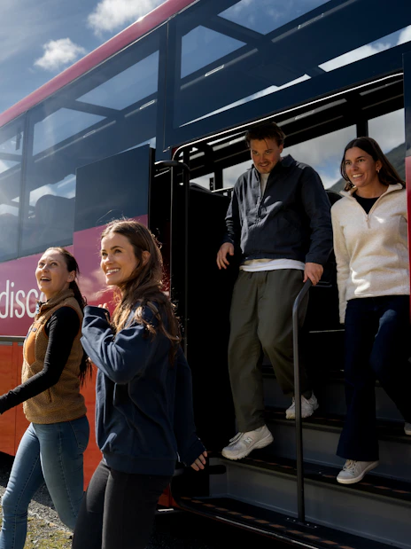 Tourists exiting a coach at Milford Sound with scenic mountain backdrop.