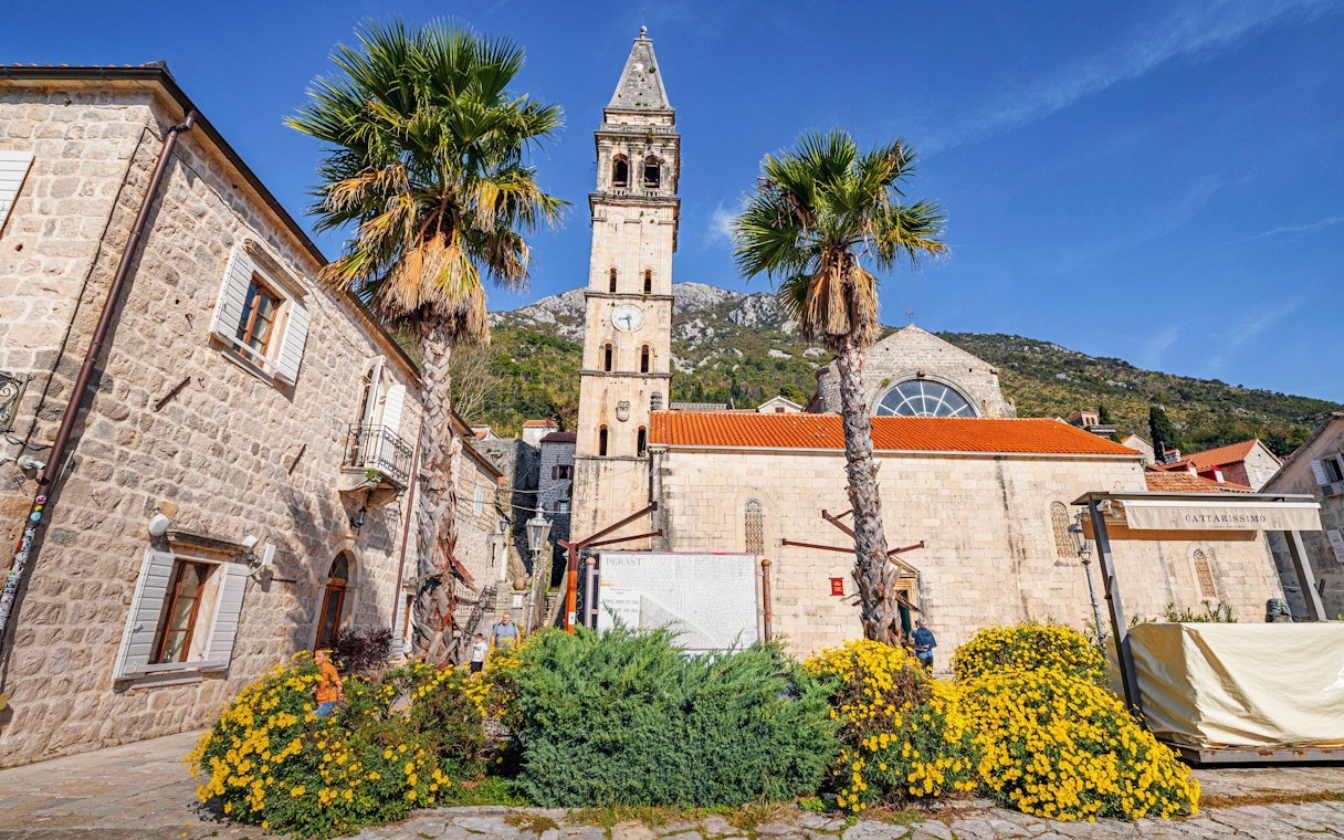 Saint Nicholas' Church with bell tower in Perast, Montenegro, surrounded by palm trees and flowers.