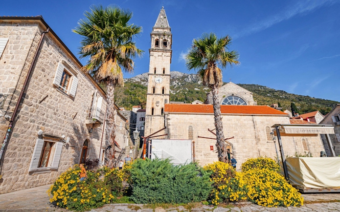 Saint Nicholas' Church with bell tower in Perast, Montenegro, surrounded by palm trees and flowers.