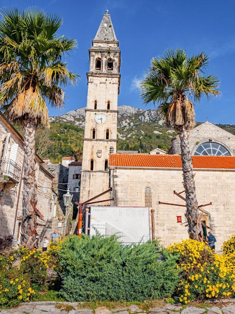 Saint Nicholas' Church with bell tower in Perast, Montenegro, surrounded by palm trees and flowers.