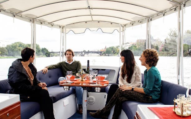 Tourists enjoying drinks and snacks on a Seville river cruise with city views.
