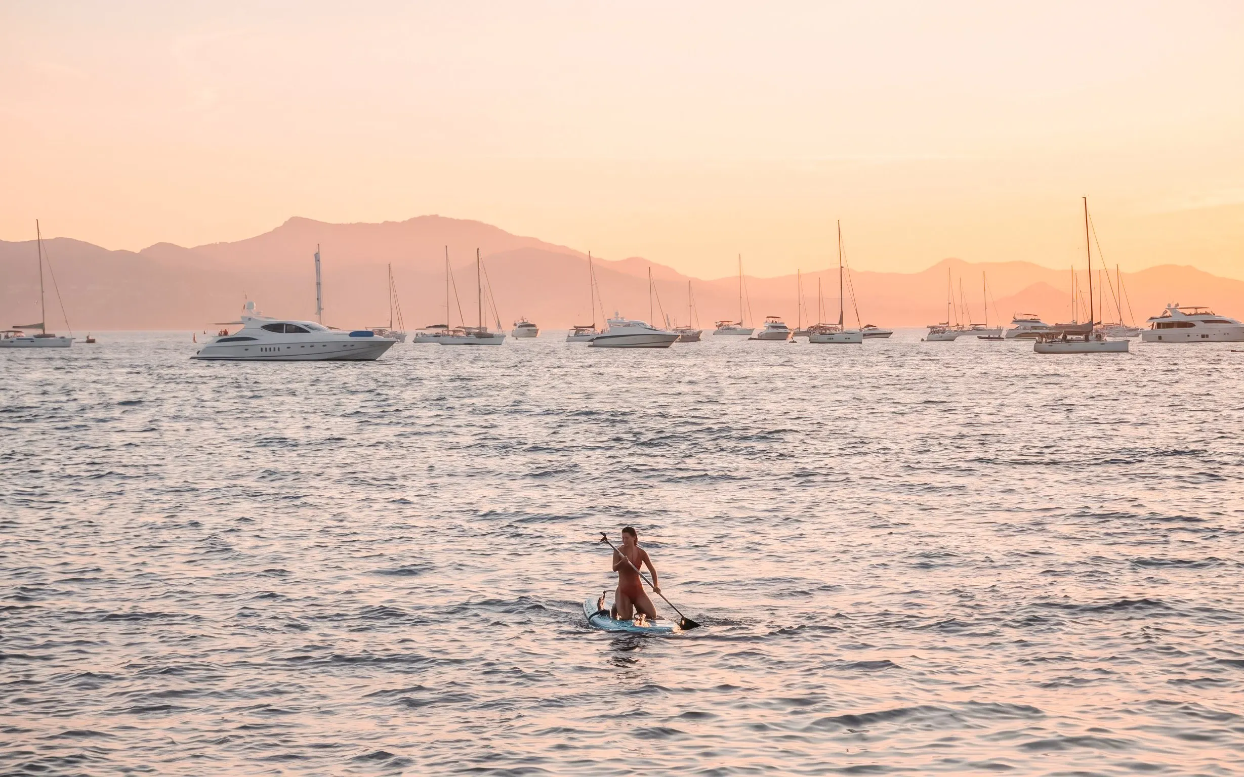 Paddleboarder on calm sea at sunset with yachts in French Riviera.