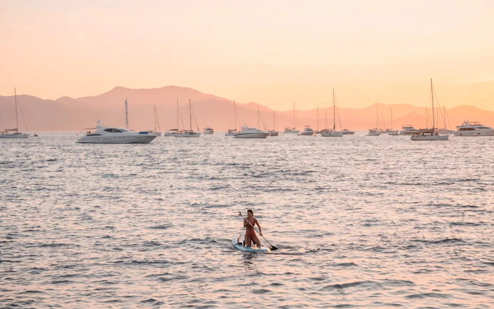 Paddleboarder on calm sea at sunset with yachts in French Riviera.