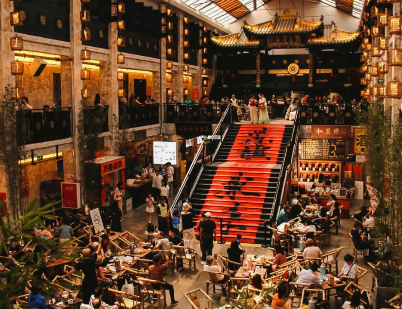 Indoor dining area with people seated at tables near a large staircase with red banners, Chengdu.