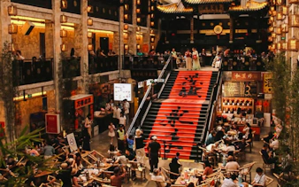Indoor dining area with people seated at tables near a large staircase with red banners, Chengdu.