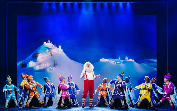 Santa and elves performing on stage in "Elf The Musical" with snowy village backdrop.