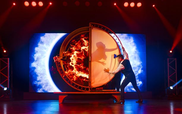 Magician performing illusion with fiery backdrop at Evolution Magic Show.