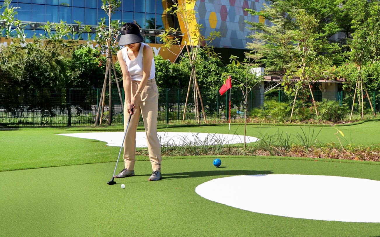 Golfer putting on a mini-golf course with modern building in the background.