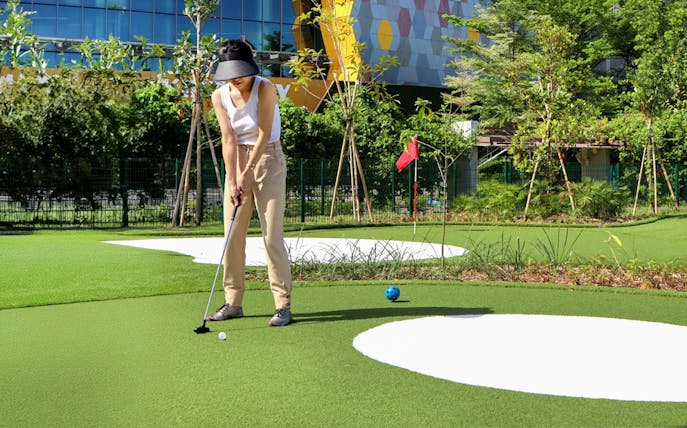 Golfer putting on a mini-golf course with modern building in the background.